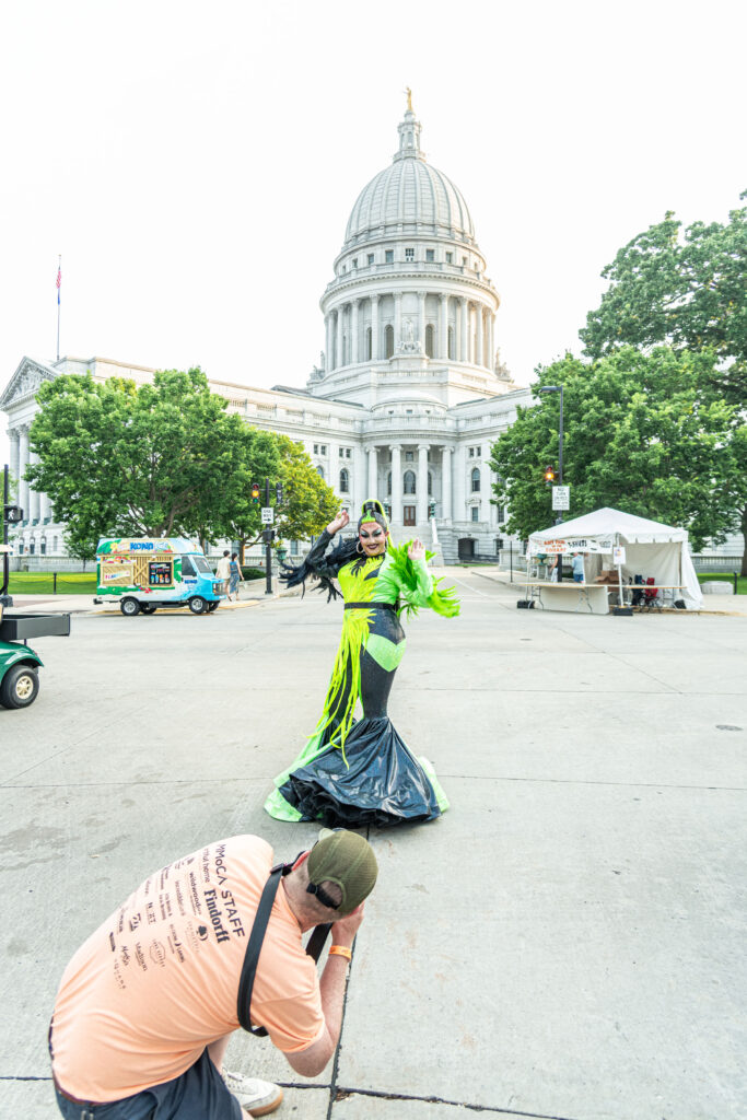 A man in an orange shirt taking photos of a drag queen in a green dress in front of the wisconsin state capitol